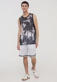 Canotta bianca e nera con design di palma, abbinata a pantaloni corti bianchi a righe e sneakers bianche con accenti marroni.