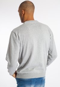 Man med kort hår som bär en ljusgrå långärmad tröja och blå jeans, vänd bort mot en enkel vit bakgrund.