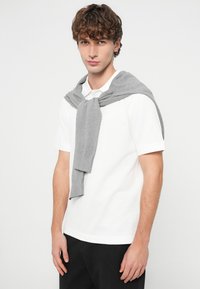 Λευκό πόλο πουκάμισο με γκρι πλεκτό πουλόβερ περασμένο πάνω από τους ώμους. Το μοντέλο στέκεται μπροστά σε απλό φόντο, παρουσιάζοντας τις λεπτομέρειες της εμφάνισης.