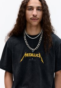 Černé tričko s žlutou grafikou "Metallica", oblečené s dvěma vrstvenými řetězy: silným stříbrným řetězem a jemným perlovým náhrdelníkem.