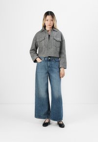 Grå, kort ulljakke med to brystlommer, kombinert med vide blå denimjeans. Modellen har på seg svarte leiligheter. Enkel bakgrunn.