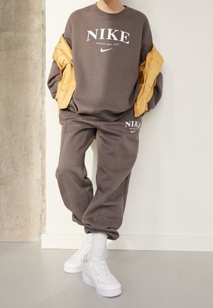 Persona che indossa felpa e pantaloni Nike marroni con sneakers bianche e una giacca gialla sulle braccia, in piedi all'interno.