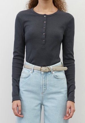 Frau trägt ein eng anliegendes, dunkelgraues, langärmliges Henley-Shirt mit Rippstruktur, das in hellblaue, hochtaillierte Jeans mit beigem Gürtel gesteckt ist.