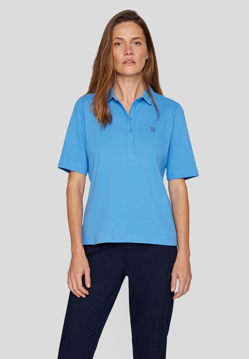 Hellblaues Kurzarm-Polohemd mit Knopfleiste und gesticktem Logo auf der Brust. Glattes Material, lässige Passform, kombiniert mit dunklen Hosen.