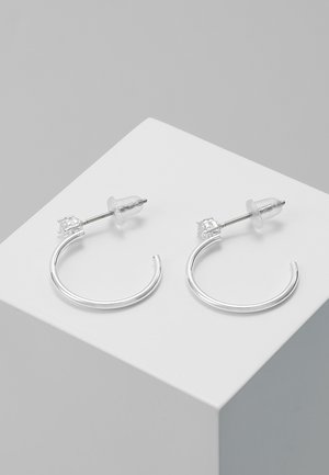 Pari pieniä, ohuita hopeisia renkaskorvakoruja, joissa kirkkaat muoviset tukihelat, esillä valkoisella geometrisella pinnalla.