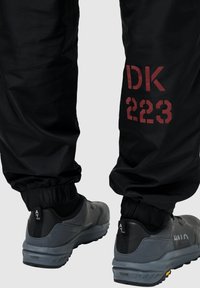 Czarne wodoodporne spodnie z elastycznymi mankietami, z czerwonym nadrukiem "DK 223". W komplecie z szarymi butami sportowymi, które prezentują teksturowany materiał i wzór.