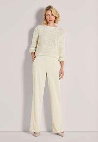 Vrouw met kort blond haar, gekleed in een crèmekleurige gebreide trui, wijdvallende crèmekleurige broek en beige hakken, staande tegen een lichte muur.