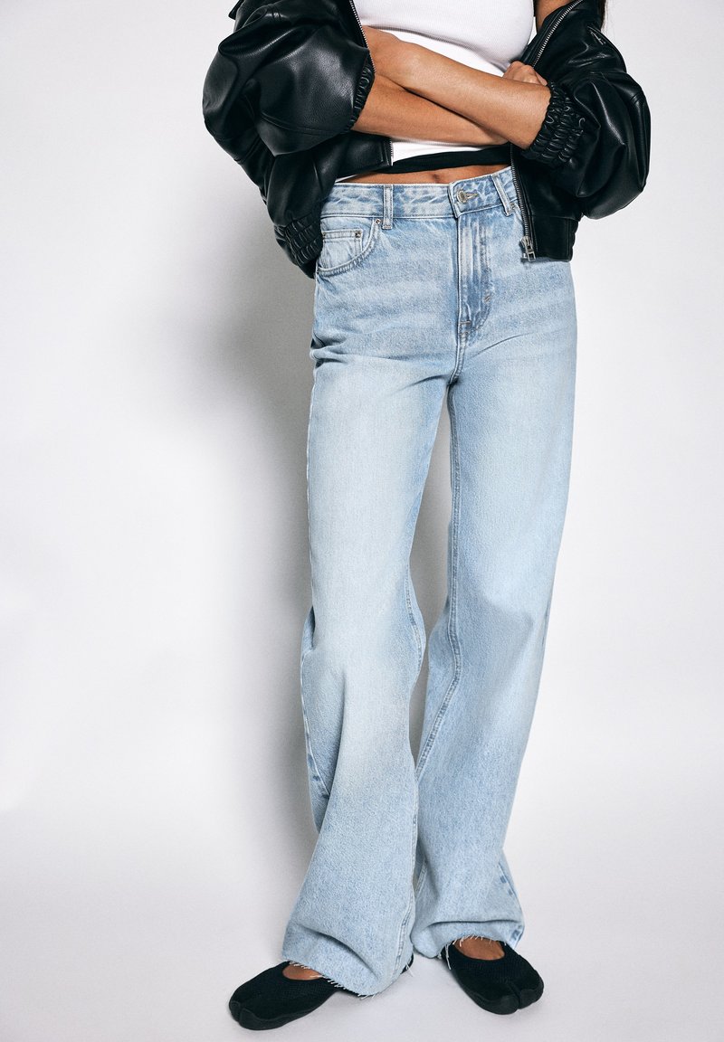 Personne portant un jean évasé bleu clair, des chaussures noires sans lacets, un débardeur blanc et une veste en cuir noire, bras croisés.