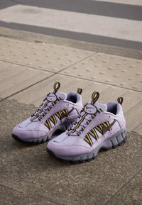 Lila sneakers med svarta och gula accenter, texturerade sulor och ett framträdande snörningssystem. Har en vadderad krage och meshpartier.