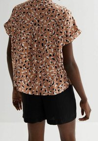 Krátký rukáv, volně střižená blůza v terakotové barvě s černobílým abstraktním vzorem. Skombinována s černými šortkami.