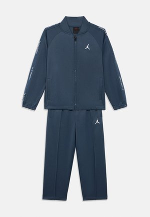 Mørkeblå atletisk treningsdress laget av mykt materiale, med en jakke med glidelås og bukser med elastisk midje, med hvite logodetaljer på begge.