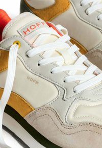 Zapatilla blanca y beige con paneles de ante y tela, acentos naranjas, cordones blancos y suela texturizada. Logotipo "HOFF" en el lateral.