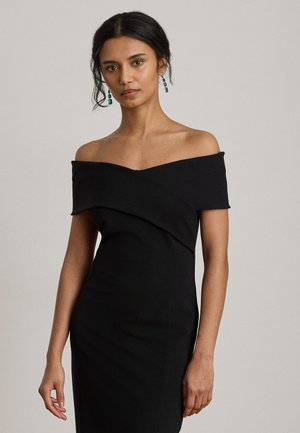 Un mannequin pose devant un fond uni, vêtu d’une robe noire à épaules dénudées et de boucles d’oreilles pendantes turquoise.