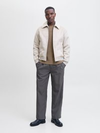 Bărbat stând cu mâinile în buzunare, purtând o geacă bej de blană, pulover maro, pantaloni gri și pantofi negri, pe fundal alb.