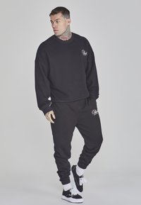 Set di felpa e pantaloni della tuta neri, con una vestibilità ampia, polsini a coste e dettaglio del logo su petto e coscia. Materiale in cotone, texture morbida.