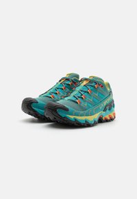 Zapatillas de trail running en verde azulado y negro, con parte superior de malla, puntera de goma, acentos en amarillo y rojo, y suela con agarre texturizado.
