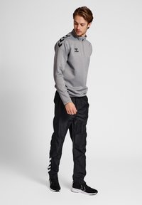 Grå långärmad zip-top med svart axelchevronmönster, tillsammans med svarta joggers med vita randaccenter och svarta sportskor.