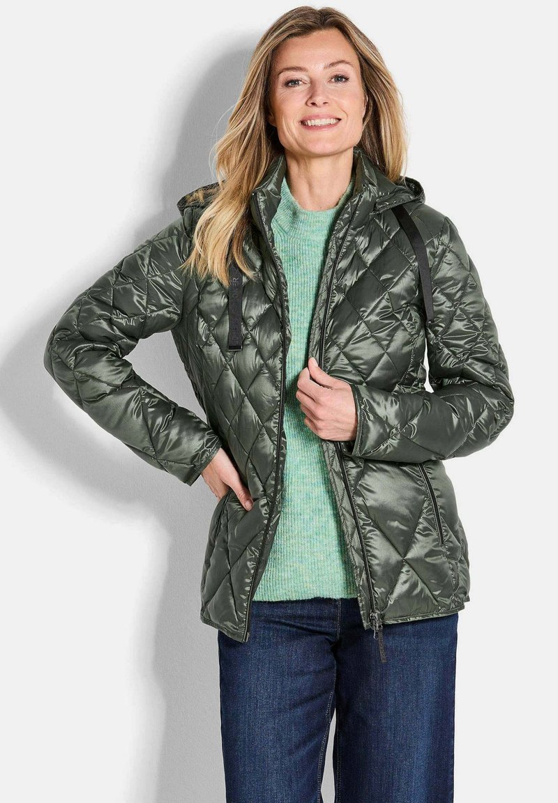 Gestepptes grünes Jacket mit glänzender Oberfläche, ausgestattet mit Kapuze, schwarzen Akzenten und Reißverschluss. getragen über einem hellgrünen Pullover und blauen Jeans.