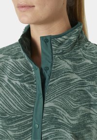 Groene fleecejack met een golfpatroon, voorzien van een knoopsluiting bij de kraag en een gladde voering. De gestructureerde buitenkant contrasteert met de effen binnenlaag.