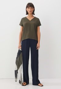 Top verde oliva a V con maniche corte, abbinato a pantaloni navy a gamba larga. Un foulard con fantasia posato su un braccio completa il look.