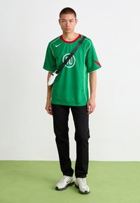 Jauns vīrietis stāv, ģērbts zaļā sporta kreklā, melnās biksēs, baltās kedās un baltā plecu somā ar melnu siksnu uz zaļa grīdas.