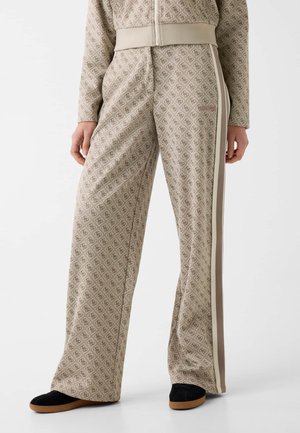 Pantalon ample à motifs beige avec des bandes latérales, porté avec un haut zippé assorti et des chaussures slip-on noires avec des semelles marron.