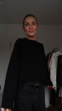 Schwarzer Pullover mit langen Ärmeln und lockerem Schnitt, kombiniert mit schwarzer Jeans und Gürtel. Im Hintergrund sind Kleidungstücke an Bügeln zu sehen.