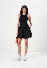 Zwart mouwloos jurkje met een geplooide rok, gemaakt van een gladde stof, gecombineerd met witte high-top sneakers en een kleine rode handtas.