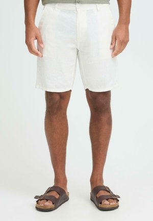 Shorts en lin blanc à coupe décontractée, dotés de poches et d'ourlets retroussés, associés à des sandales marron avec des détails visibles des pieds.