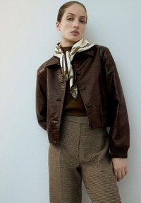 Mujer vestida con una chaqueta de cuero marrón texturizado, bufanda de seda estampada, top marrón y pantalones de pata de gallo de talle alto contra un fondo neutro.