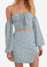 Haut court bleu à fleurs, épaules dénudées, devant froncé et manches longues bouffantes, assorti d'une jupe fendue sur le côté.