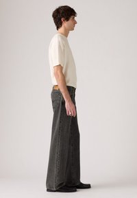 Man in een crèmekleurig T-shirt, ruime zwarte jeans en zwarte schoenen die zijwaarts staat tegen een effen achtergrond.