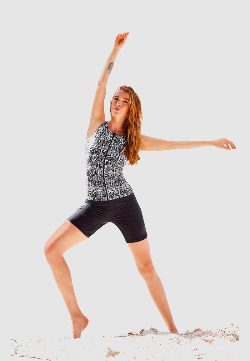 Zwart-wit gestreepte atletische jumpsuit met een ritssluiting aan de voorkant, gedragen met zwarte fietsshorts. Model poseert met opgetilde armen.
