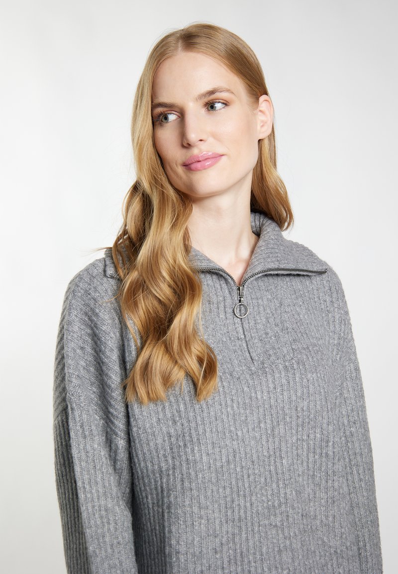 Junge Frau mit langen blonden Haaren, die einen grau gerippten Pullover mit Reißverschlusskragen trägt, seitlich gegen einen schlichten hellen Hintergrund blickt.