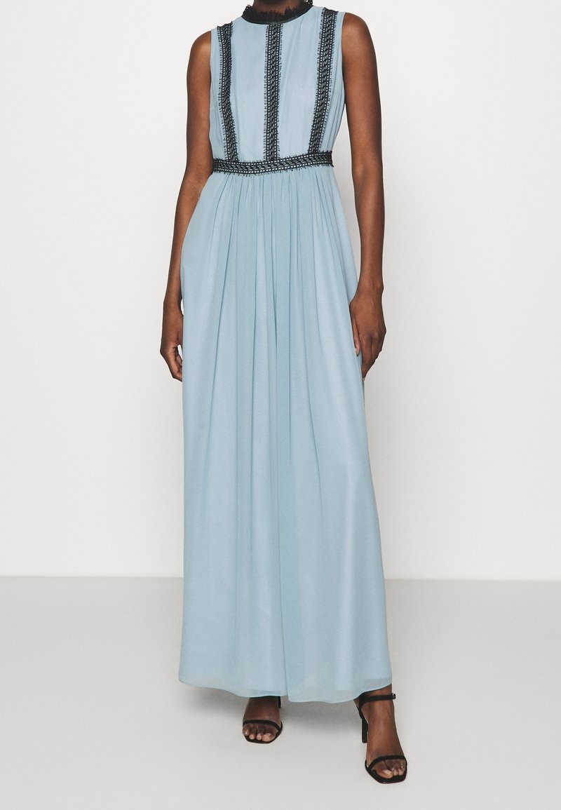 Femme portant une robe longue sans manches bleu clair avec une bordure verticale noire et des sandales à talons noires, debout sur fond blanc.