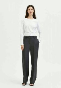Woman standing wearing a white long-sleeve shirt, dark gray wide-leg pants, and black open-toe sandals against a plain background.