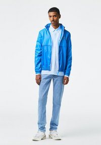 Giacca leggera blu con zip e cappuccio, abbinata a una camicia button-up azzurro chiaro, jeans azzurro chiaro e sneakers bianche.