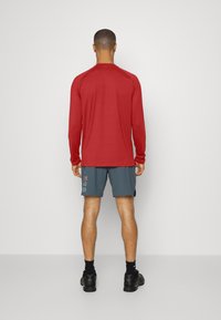 Langärmliges rotes Performance-Shirt kombiniert mit grauen Shorts, die mit numerischem Text versehen sind. Das Modell trägt schwarze Sneaker und Socken.