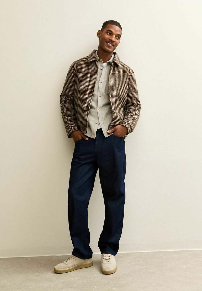Chaqueta de pata de gallo marrón con una camisa debajo, combinada con pantalones de color azul oscuro y zapatillas de color claro. De pie contra una pared neutra.
