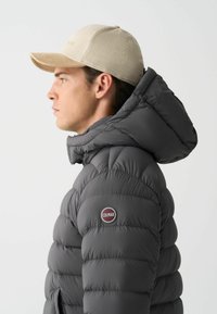 Grå pufferjakke med en aftagelig hætte, udstyret med et rundt Colmar-logo på brystet. Sammen med en beige corduroy-kasket.