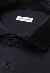 Camicia nera con colletto strutturato, dotata di un'etichetta bianca con la scritta "seidensticker". Dettagli con bottoni in un colore scuro coordinato. Tessuto liscio.