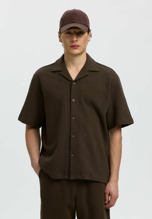 Młody mężczyzna w ciemnobrązowej, krótkiej koszuli zapinanej na guziki, do tego dopasowane spodnie oraz brązowa czapka z daszkiem, stojący na jednolitym tle.