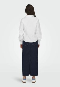 Camicia bianca a maniche lunghe con colletto, abbinata a una lunga gonna di jeans scuri e sneakers bianche con dettagli blu, vista da dietro.