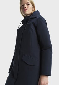 Mlada žena nosi tamnoplavi zimski kaput s kapuljačom i patentnim džepovima, malo gleda prema gore na svijetloj pozadini.