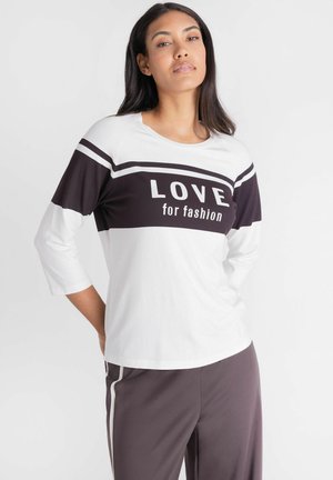 Vrouw staand met handen achter rug, draagt witte en zwarte top met tekst "LOVE for fashion" en donkere broek met witte strepen.