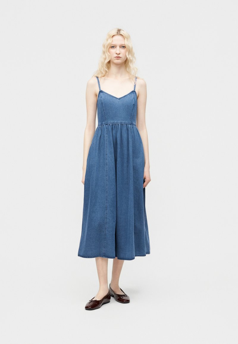 Jeune femme debout portant une robe midi en denim bleu sans manches avec fines bretelles et des ballerines rouge foncé sur un fond uni.