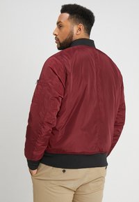 Giacca bomber rossa con finitura liscia, colletto e polsini neri a coste, tasca con zip sulla manica sinistra e vita elasticizzata.