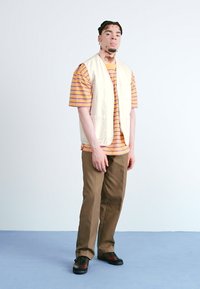 Beige ujjatlan mellény egy színes csíkozott póló felett, barna nadrág és fekete piros akcentussal ellátott cipő. Egyszerű, laza dizájn.