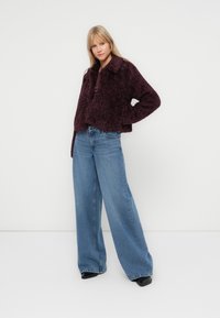 Bordeaux fuzzy cropped jack met een kraag, gecombineerd met wijde lichtblauwe jeans. De jack heeft een zachte textuur en een knoopsluiting.