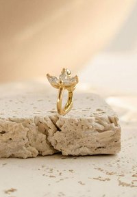 Anello d'oro con un design floreale che presenta tre gemme trasparenti tagliate a marquise, montato su una fascia texturizzata, esposto su una superficie di pietra chiara.
